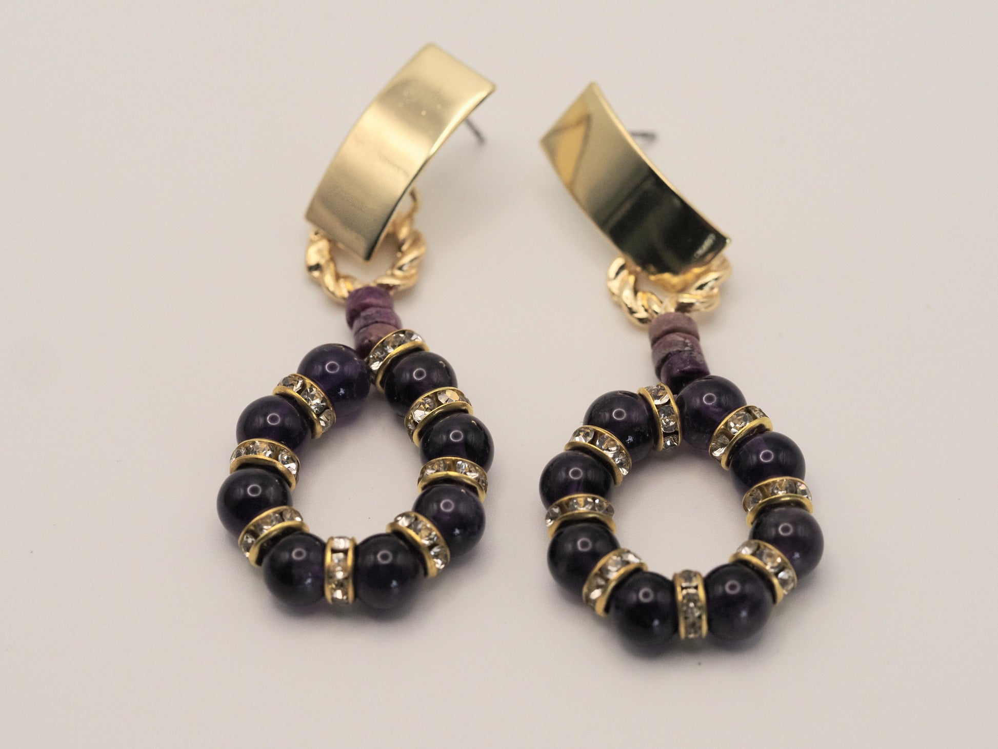 A pair of gold and dark purple beaded hoop earrings on a light gray background.